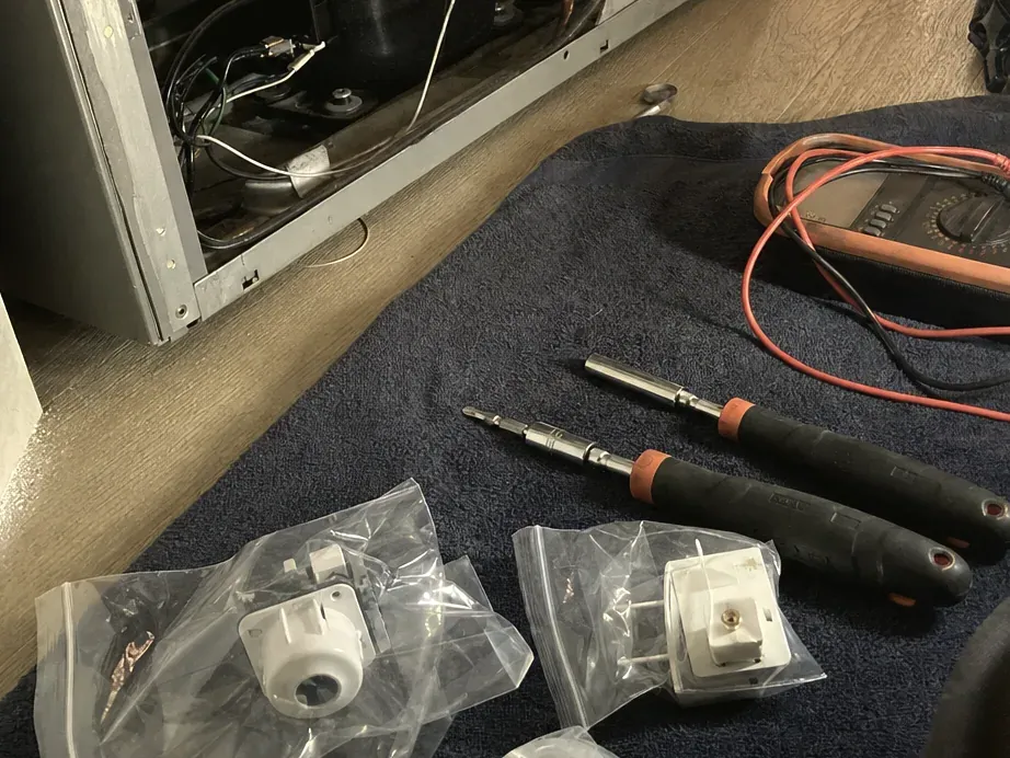 Replacement parts in clear bags beside two screwdrivers and a multimeter on a navy pad next to the open unit.