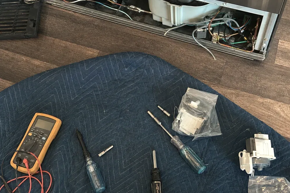 Yellow multimeter, screwdrivers and bagged replacement parts on a blue moving pad in front of an opened refrigerator base.