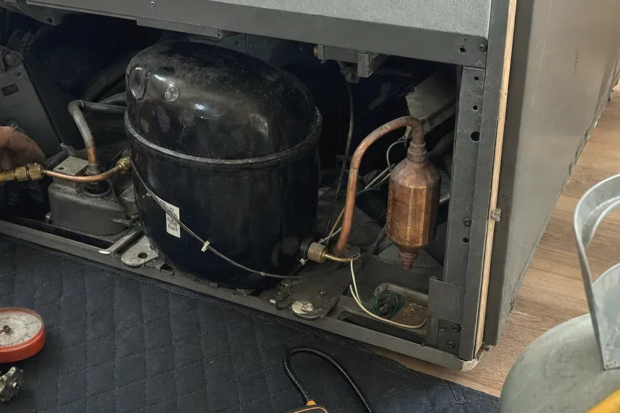 Black compressor and copper filter-drier in an open machine compartment, a refrigerant gauge on the pad below.