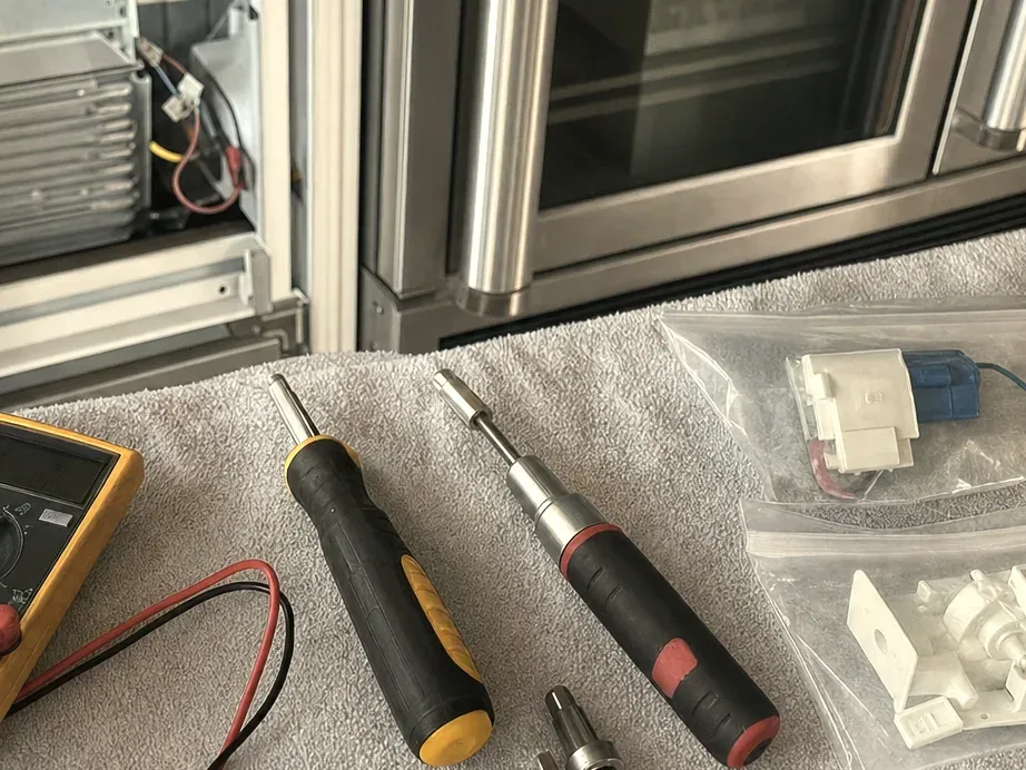 Screwdrivers, a multimeter and bagged parts on a grey towel in front of an open stainless refrigerator base.